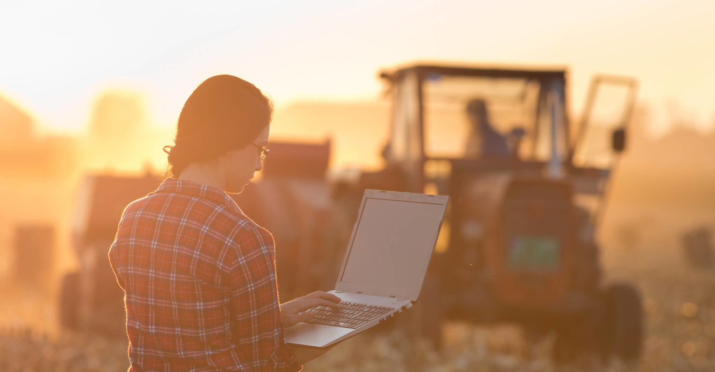 Agriculture student with laptop surveying farmers ploughing in field